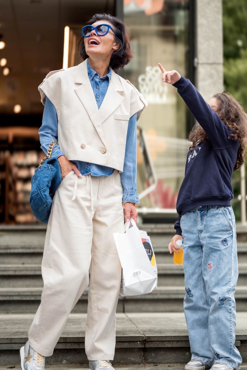 Two people on a city street, one in a white vest and beige pants, the other in a dark jacket and light jeans.