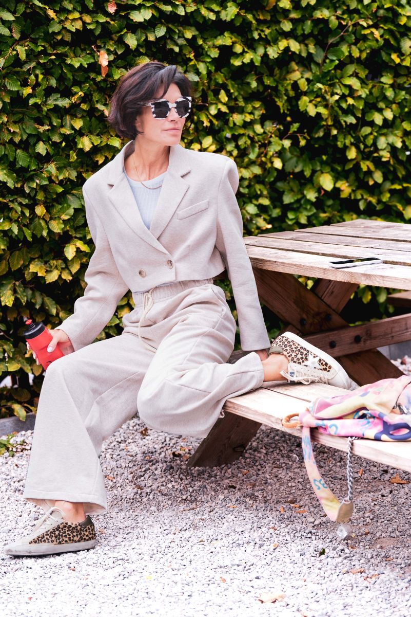 Woman in a beige suit sitting on a bench with a green hedge background