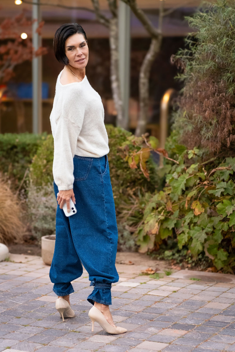 Woman wearing a white sweater and blue jeans standing outdoors on a paved path.