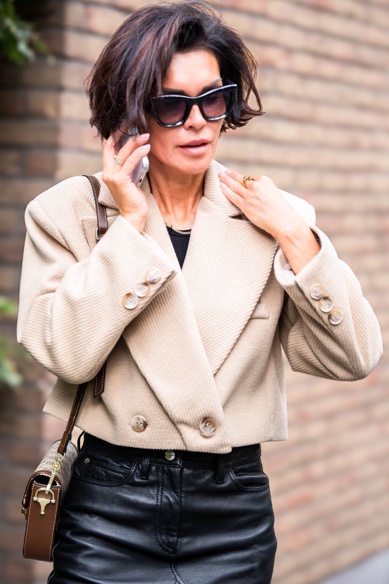 Woman wearing a beige coat and black leather pants, standing against a brick wall.