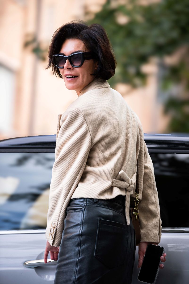 Woman in a beige jacket and black pants standing next to a car outdoors.