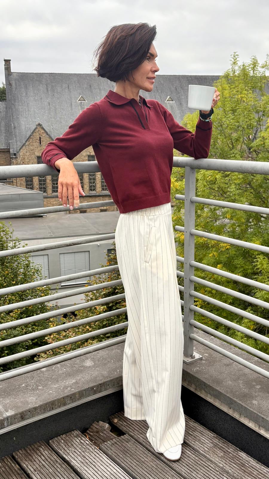 Woman standing on a balcony wearing a red top and white pants, holding a cup.