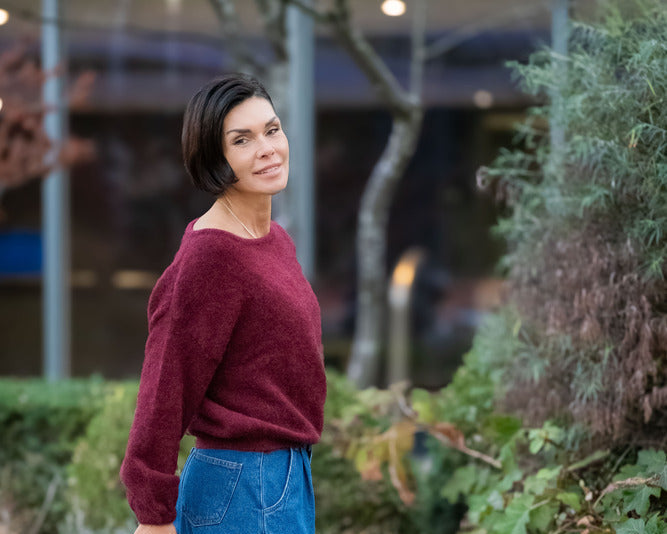 Woman in a red sweater and blue jeans standing outdoors on a paved path with greenery.