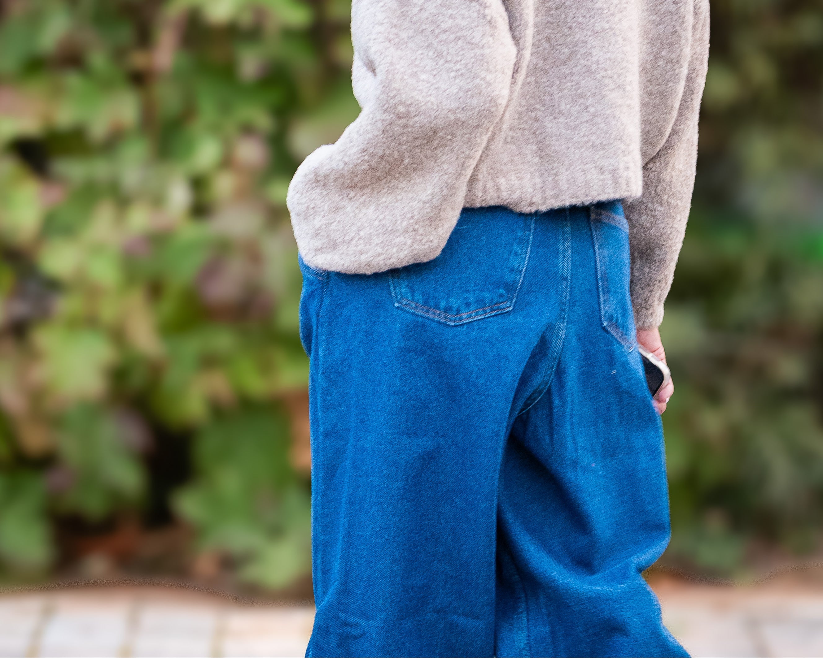 Person wearing a beige sweater and blue pants standing in front of green foliage