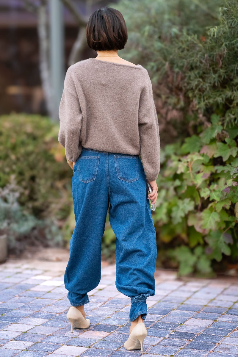 Person wearing a beige sweater and blue jeans walking on a paved path with greenery in the background