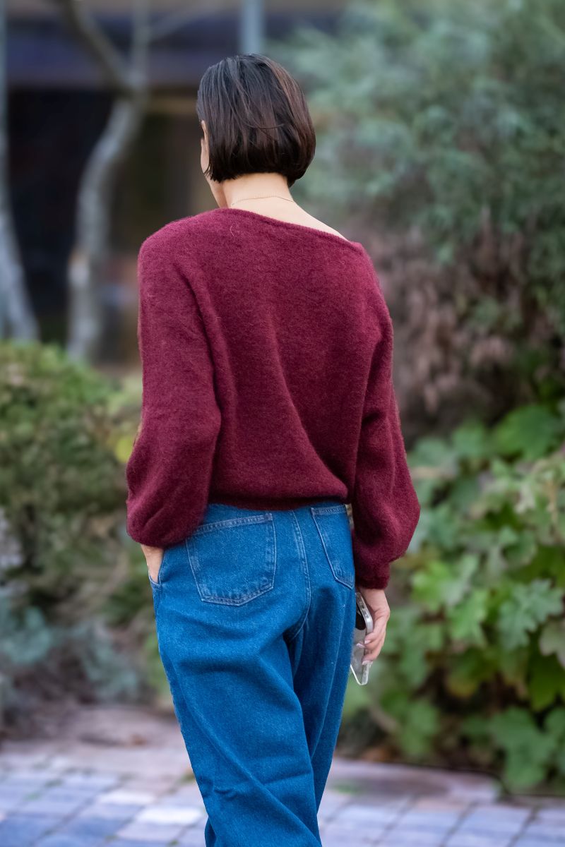 Person wearing a burgundy sweater and blue jeans standing outdoors with greenery in the background