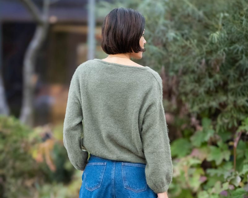Person wearing a green sweater and blue jeans walking on a paved path with greenery in the background