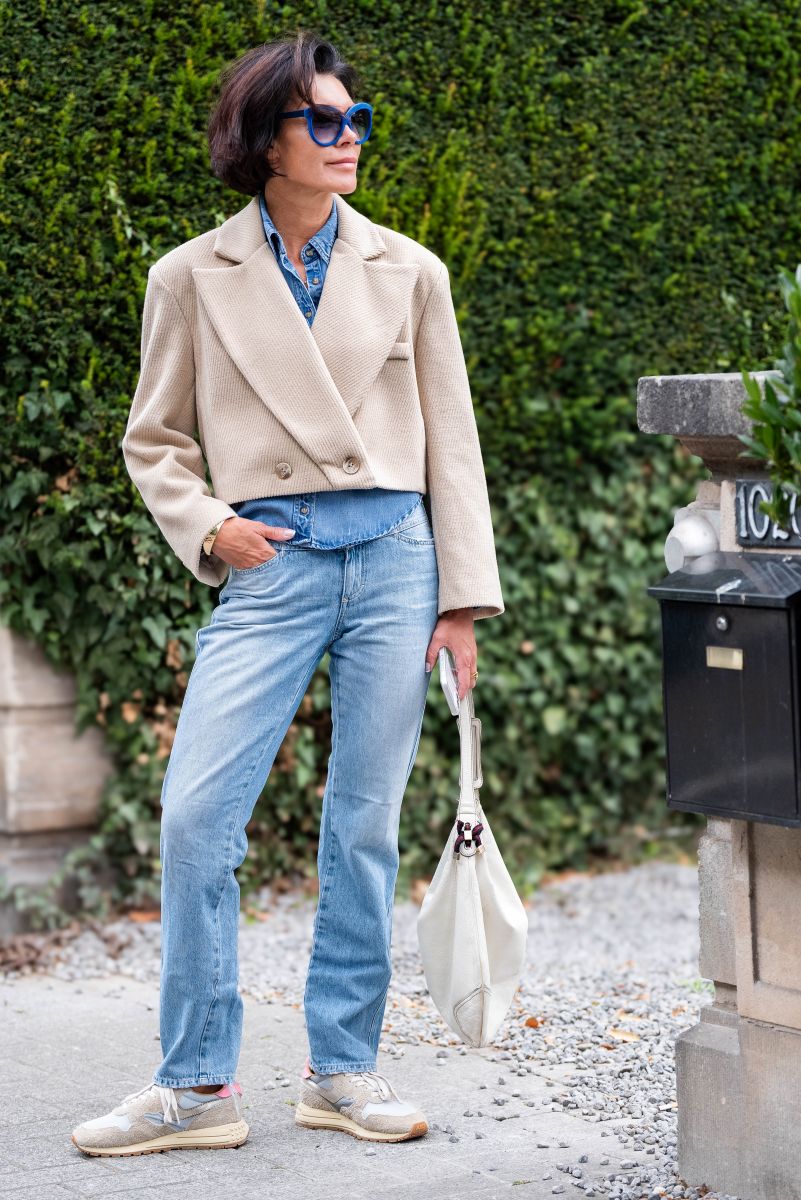 Person wearing a beige coat, blue jeans, and sunglasses standing outdoors with greenery in the background.