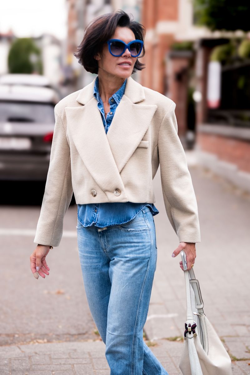 Woman walking on a street wearing a beige jacket, blue jeans, and sunglasses.