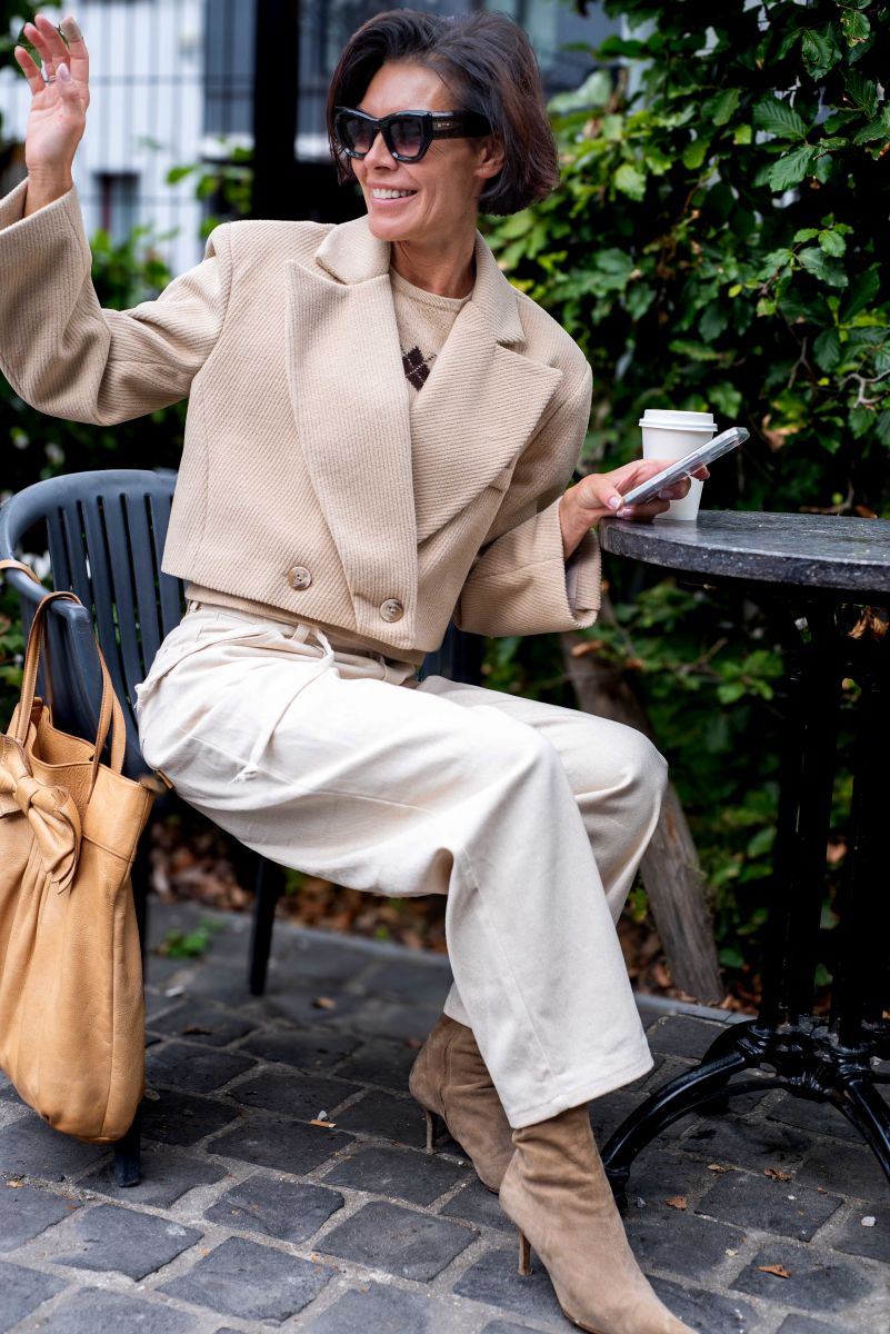 Woman in beige coat and sunglasses sitting outdoors, holding a phone and a cup.